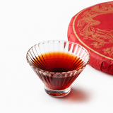 Brewed Rainforest Ancient Tree Ripe Pu-erh tea in a clear glass cup with red tea cake packaging on a white background