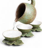 Speckled green ceramic tea pitcher pouring water into matching handmade teacups on a clean white background
