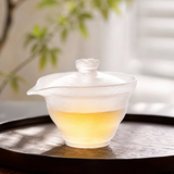 Frosted glass gaiwan filled with golden tea on a tea tray in soft natural light, highlighting the translucent glass texture and premium tea brewing style.