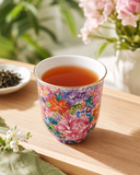 Floral porcelain aroma cup filled with amber black tea, placed on a clean wooden tea table with soft green plants and pink flowers in the background.